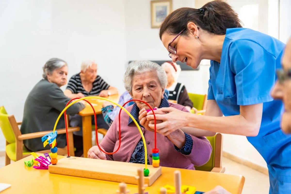 Caregiver assists elderly woman with a bead maze activity in a bright room